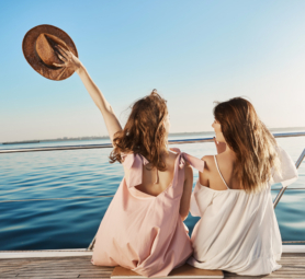 Back portrait of two female friends sitting on boat, waving with hat while talking and enjoying looking at seaside. Sisters finally took vacation to visit their mom who lives in Italy.